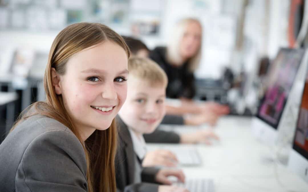 Students from Laurus Cheadle Hulme work on computers with the support of a teacher, out of focus in the background. A girl in the foreground, in focus, smiles.