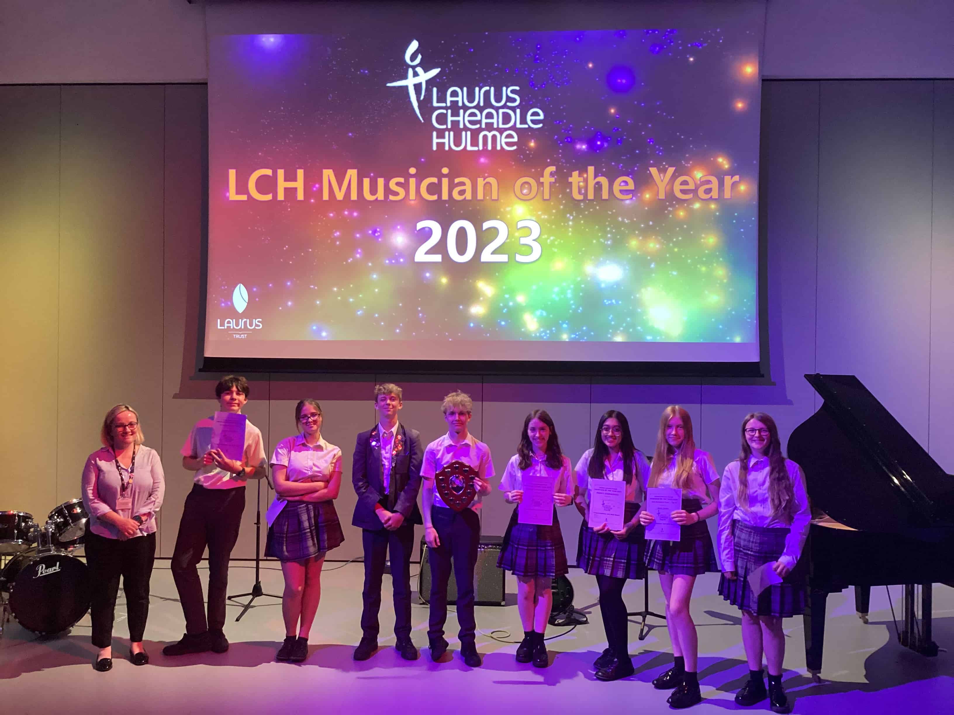 The 8 nominated performers in the Laurus Cheadle Hulme Musician of the Year competition 2023 stand next to Miss Brown (Trust Director of Music) on the stage in purple stage lighting. Behind them, a projector screen says LCH Musician of the Year 2023.