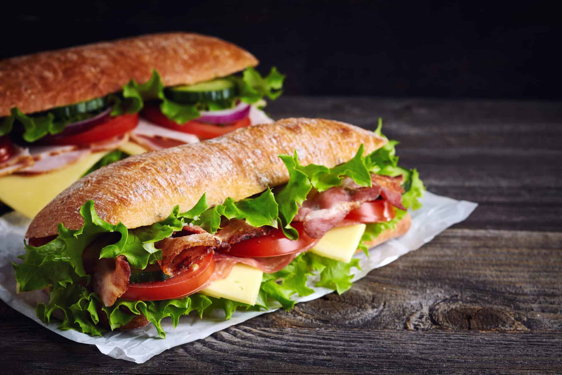 An example of food provided by Dolce at Laurus Cheadle Hulme canteen. Two fresh submarine sandwiches with ham, cheese, bacon, tomatoes, lettuce, cucumbers and onions on dark wooden background